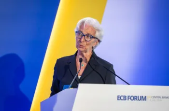 La presidente della Bce, Christine Lagarde, durante un intervento. Davanti a lei un podio con microfono, dietro uno sfondo con i colori blu e giallo alternati. Indossa una giacca scollata taiileur e occhiali neri