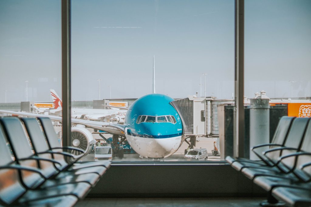 Dall'interno della sala d'attesa di un'aerostazione si vede un aereo con il colore turchese di KLM con il tunnel per i passeggeri collegato