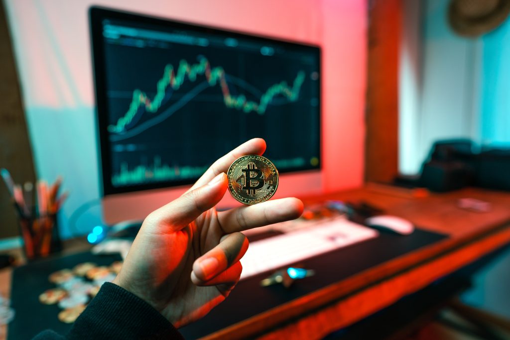 Una mano tiene tra due dita un Bitcoin mentre sullo sfondo uno schermo di pc mostra l'andamento di una attività finanziaria
