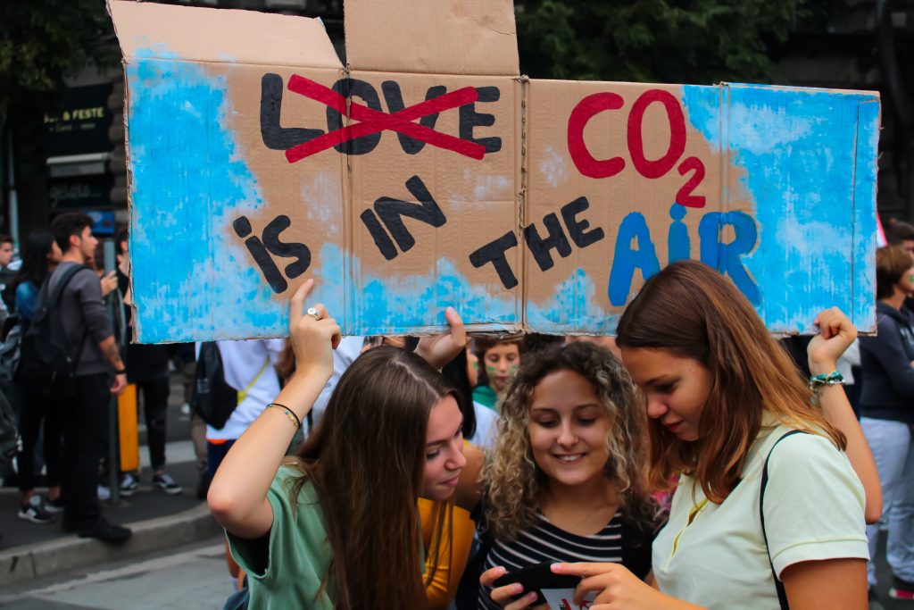 Tre ragazze guardano lo schermo di uno smartphone nel corso di una manifestazione contro l'inquinamento. Sullo sfondo un cartello con la parola LOVE sostituita da quella CO2 is in the air