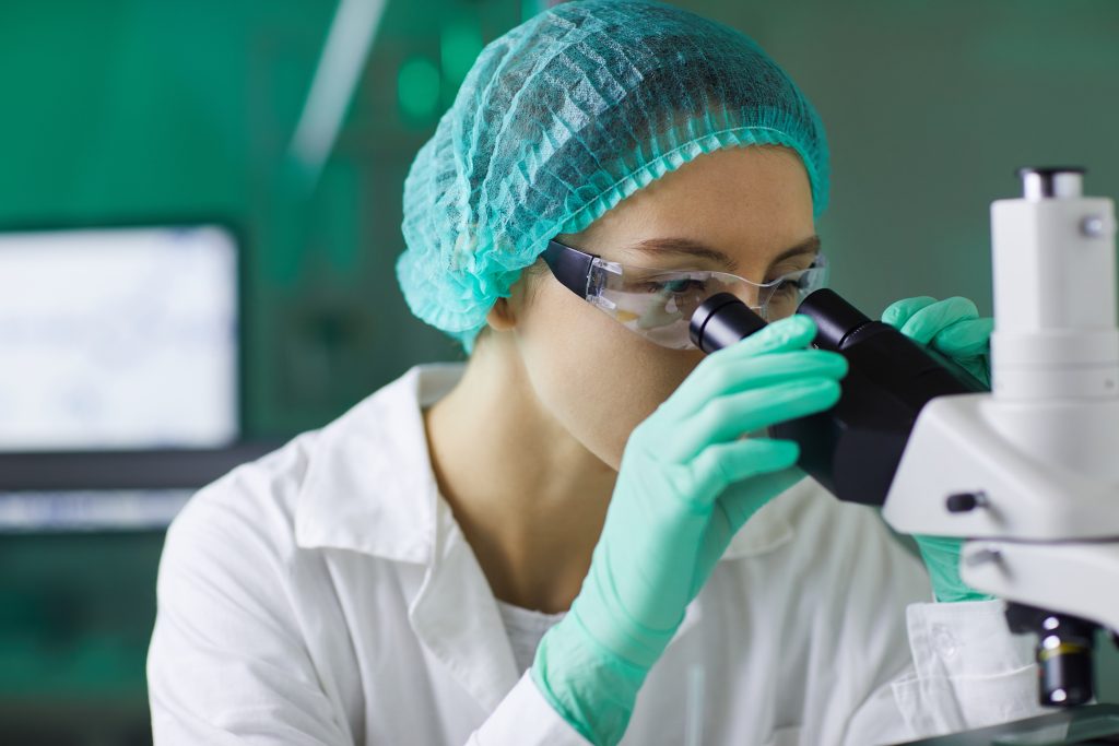 Una ricercatrice in camice bianco e guanti e cuffia verdi effettua delle osservazioni al microscopio