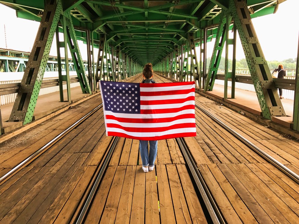 Una ragazza, di spalle, cammina su un ponte ferroviario coperto con la bandiera statunitense sulle spalle. Foto di volligda da Envato