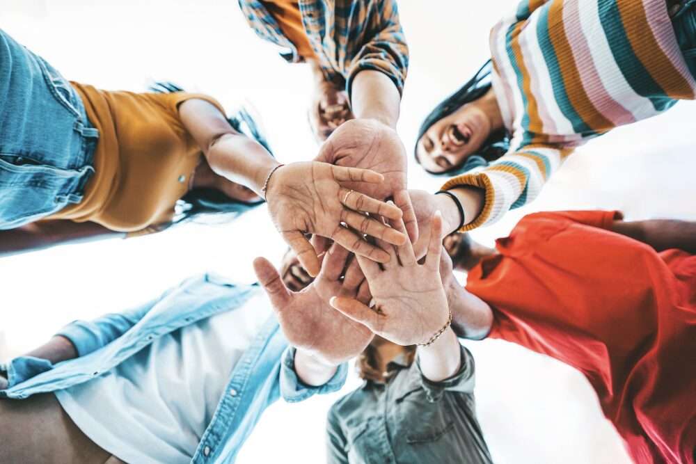Un gruppo di sei giovani con abiti colorati, posti in cerchio, unisce le mani al centro. La foto è scattata dal basso