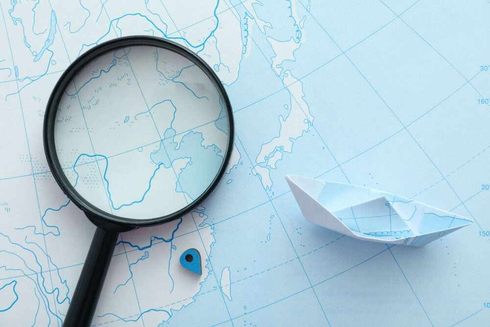 Nell'immagine, una lente d'ingrandimento e una barchetta di carta sono poggiate su una mappa in corrispondenza delle coste di Cina e Giappone