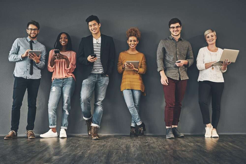 L'immagine mostra un gruppo di sei millennials diversamente vestiti e con in mano tablet, laptop o telefonini, poggiati con le spalle su un muro grigio scuro