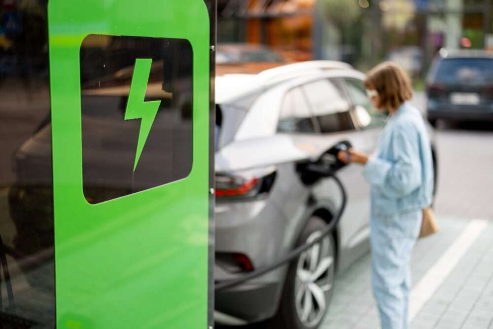 Nell'immagine, una colonnina per la ricarica elettrica degli autoveicoli e sullo sfondo, leggermente sfuocate, una donna che ricarica la sua auto