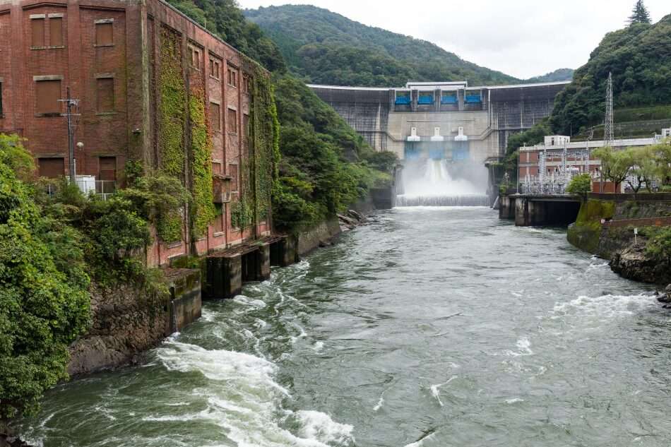 L'immagine mostra una centrale idroelettrica vista da valle. Sul fonoo la diga mostra un rilascio di acqua