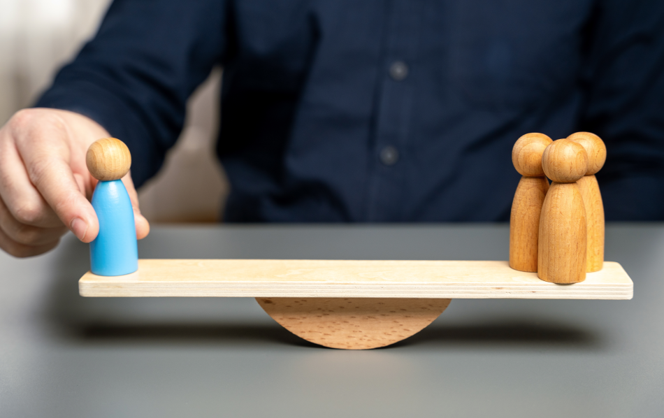 Una persona, di cui sono visibili solo le mani, riequilibra un asse di legno centrata su un fulcro poggiandovi sopra una pedina.