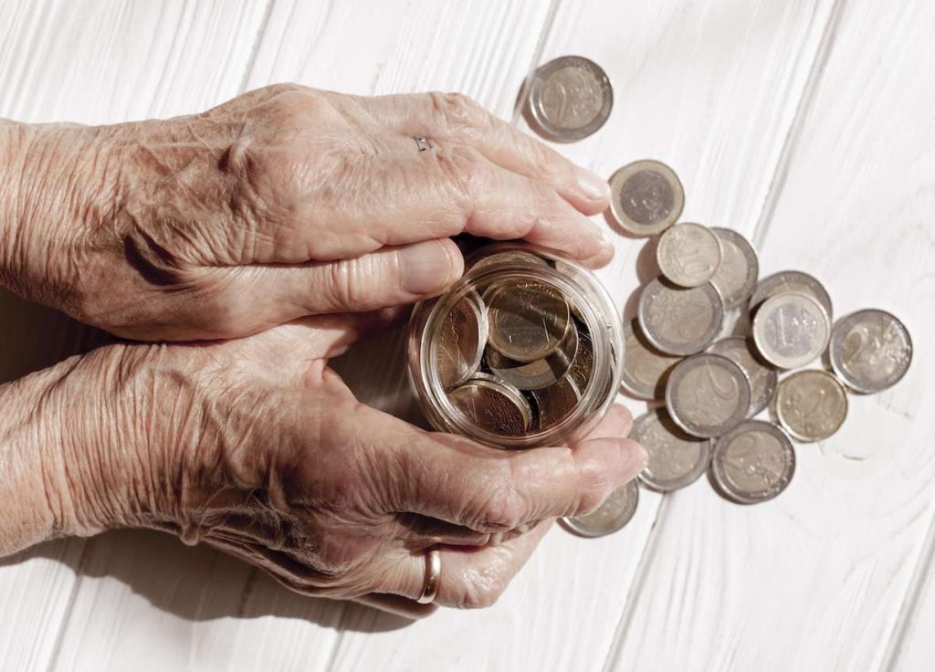 Mani di una persona anziana che tengono un barattolo di monete euro, con altre monete sparse su un tavolo bianco. Immagine simbolo di risparmi, ritiro delle pensioni di novembre alle poste