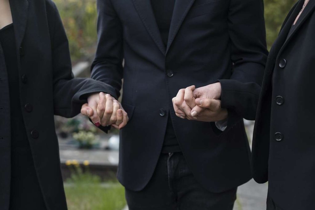 Persone in abiti scuri che si tengono per mano in segno di sostegno durante un funerale. Immagine rappresentativa di solidarietà e supporto emotivo in un contesto di lutto. Concetti correlati: spese funebri, detrazione fiscale, dichiarazione dei redditi, agevolazioni fiscali per funerali.
