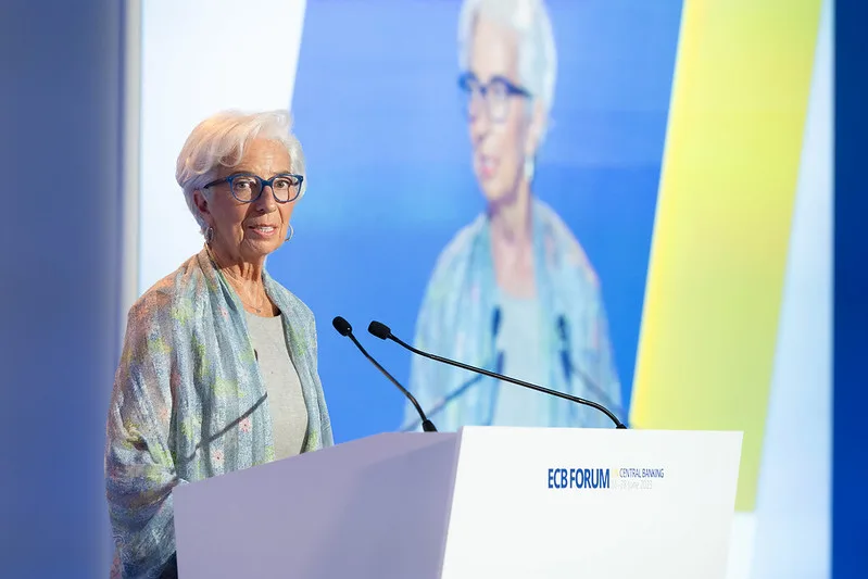 Christine Lagarde nel corso di una conferenza stampa. La fotografia, scattata lateralmente, la ritrae dietro un leggio bianco con due microfoni, vestita con una camicia azzurra aperta su una maglia grigia e occhiali scuri