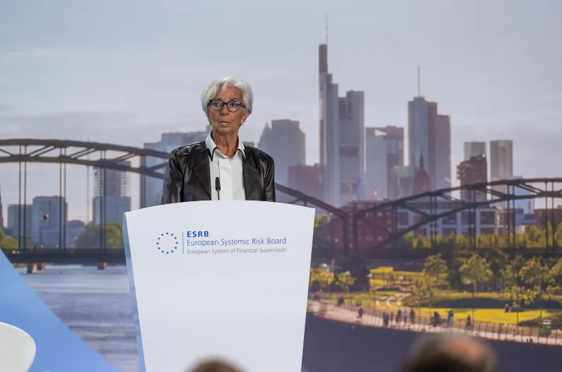 La presidente della BCE, Christine Lagarde, nel corso di un intervento al Systemic Risk Board dell'eurozona. Dietro a un leggio e davanti a uno sfondo in cui si vede la città di Francoforte con l'area dei grattacieli e il fiume Meno, indossa una giacca grigia con maglia bianca e occhiali scuri