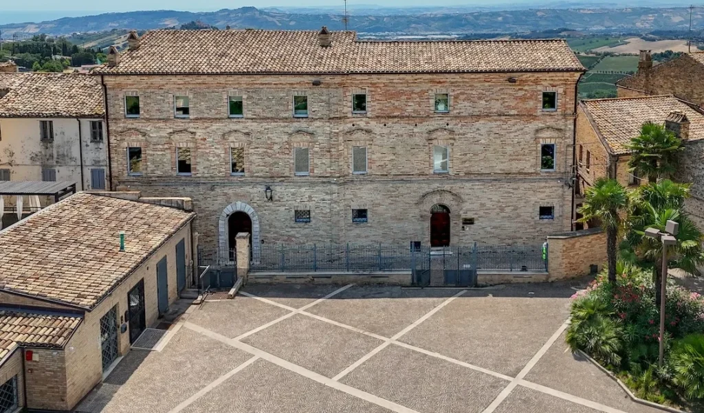 Facciata storica del Palazzo Sciarra, situato nel Piceno, con architettura in mattoni a vista e tetti in tegole. In primo piano una piazza pavimentata con motivi geometrici e vegetazione curata, sullo sfondo colline panoramiche tipiche del paesaggio marchigiano