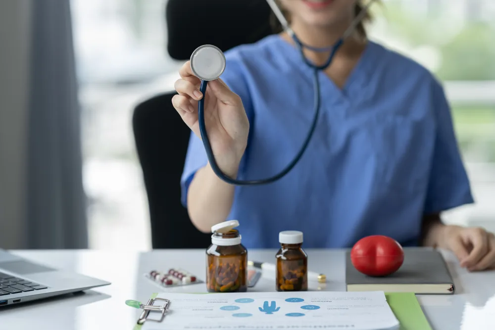 edico in camice blu con stetoscopio in mano, mostrando lo strumento verso la telecamera. Sul tavolo sono presenti flaconi di medicinali, pillole, documenti medici e un computer portatile. Ambiente medico moderno e professionale.