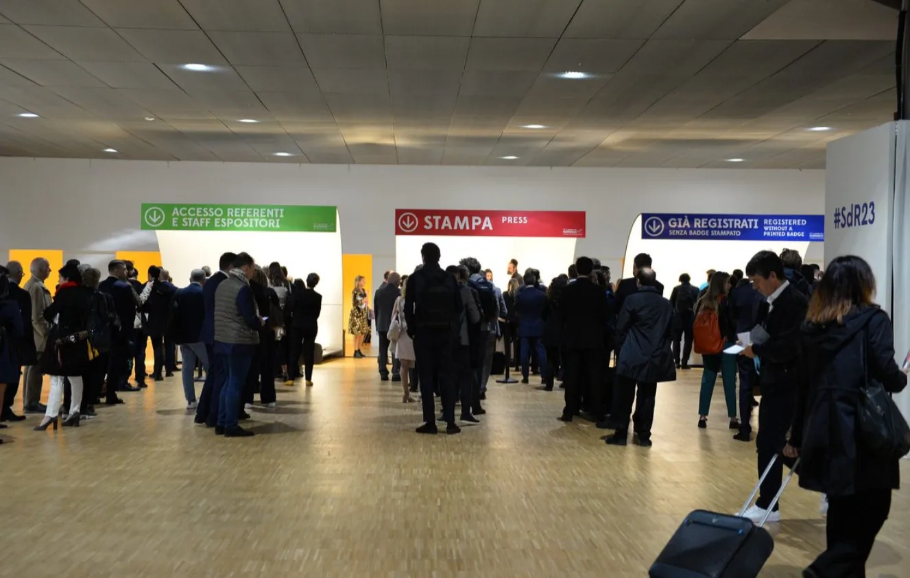 Un'immagine della hall di ingresso del Salone del Risparmio 2024. Sullo sfondo si vedono i punti di registrazione con colori differenti, verde, rosso e blu, e file di persone in attesa