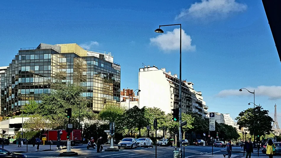 Sede centrale di Amundi a Parigi con facciata in vetro riflettente, circondata da edifici moderni e alberi, con il traffico cittadino in primo piano. Sullo sfondo, la Torre Eiffel visibile sotto un cielo sereno.
