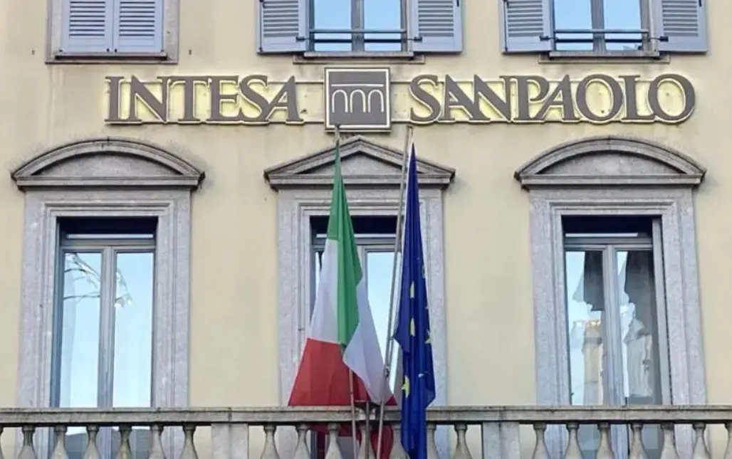 Facciata della sede di Intesa Sanpaolo con il logo dorato dell'istituto bancario italiano in evidenza. Sono visibili due bandiere, quella italiana e quella dell'Unione Europea, posizionate davanti alle finestre dello storico edificio.