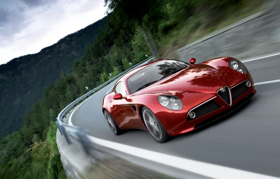 Auto sportiva Alfa Romeo di colore rosso, modello iconico in movimento su una strada di montagna con curve, circondata da vegetazione e sfondo naturale, ideale per performance di guida dinamica. Foto rappresentativa del marchio Alfa Romeo e del design italiano.