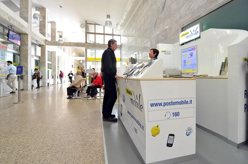 Un ufficio postale, molto grande e luminoso. Al banco, un cliente