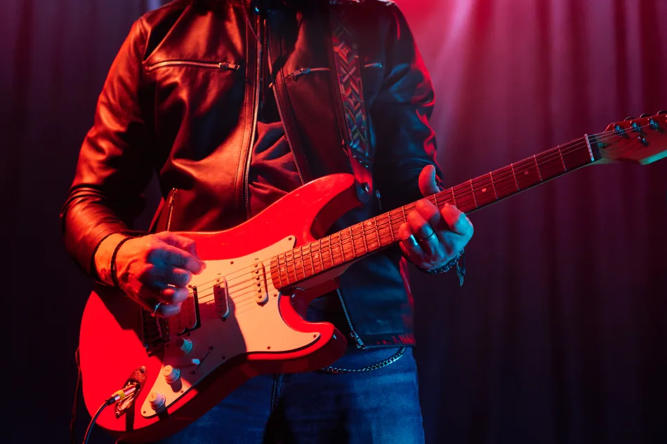 Immagine ravvicnata di un chitarrista durante un concerto rock. Nell'immagine si vede la chitarra rossa con inserto bianco. Il chitarrista indossa jeans e una giacca di pelle