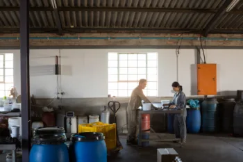 Due operai sono al lavoro all'interno di un capannone industriale illuminato da una finestra ampia sullo sfondo. Stanno controllando dei bidoni contenenti olio di oliva