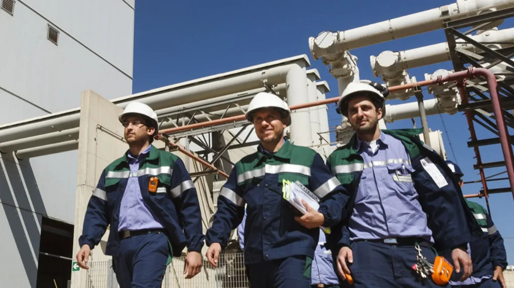 Tre ingegneri con casco protettivo e abbigliamento da lavoro camminano in un impianto industriale all'aperto, circondati da strutture e tubature, sotto un cielo sereno.