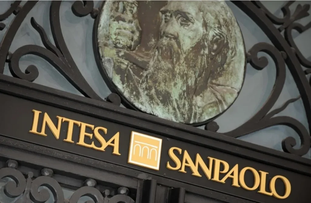 Dettaglio dell'insegna Intesa Sanpaolo con logo dorato su sfondo scuro, incorniciata da elementi decorativi in ferro battuto e rilievo artistico di figura storica.