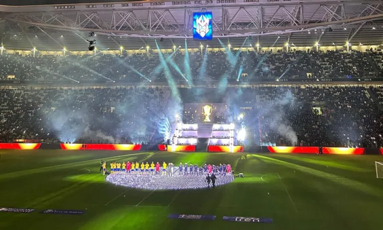 Sul prato di uno stadio di calcio, dove sono visibili anche le curve, due squadre durante la presetnazione al centro del campo: il Brasile in magilia verdeoro e la Colombia in maglia blu