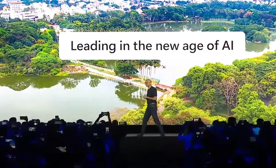 L'amministratore delegato di Microsoft, Satya Nadella, sul palco di una sala convegni durante il suo tour a Bangalore in India. Veste pantaloni scuri e maglietta nera a maniche corte. Sullo sfondo la proiezione di un panorama marino con la scritta "Leading in the new age of IA"