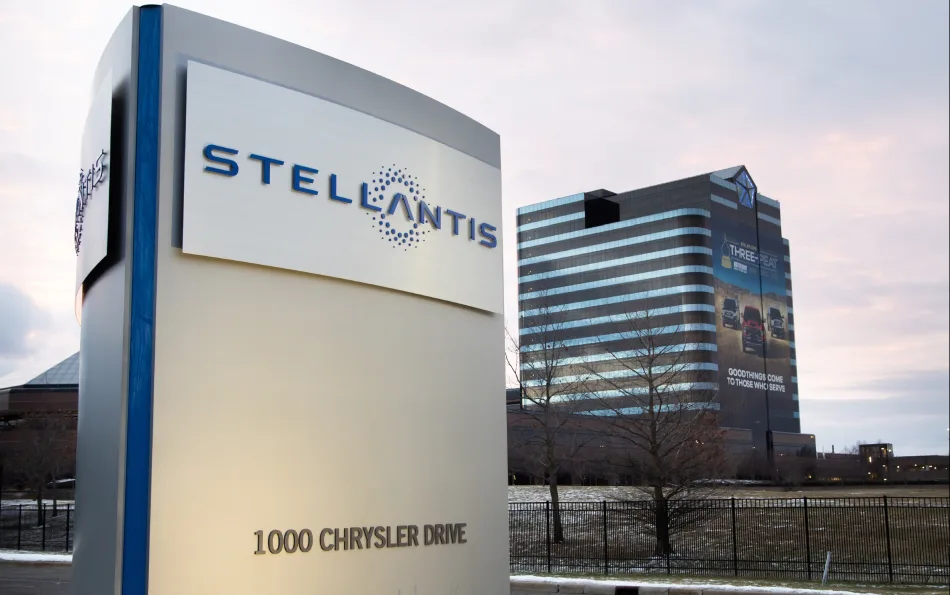 Insegna Stellantis con indirizzo '1000 Chrysler Drive' in primo piano, con la sede aziendale di Auburn Hills, Michigan, sullo sfondo al tramonto