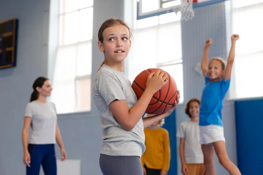 attività extra scolastiche come lo sport tra le agevolazioni del bonus studenti. Nell'immagine, all'interno di una palestra, quattro bambine giocano a pallacanestro. Una di essere ha la palla in mano e guarda alle sue spalle mentre un'altra, sullo sfondo, salta per impedire il passaggio del pallone