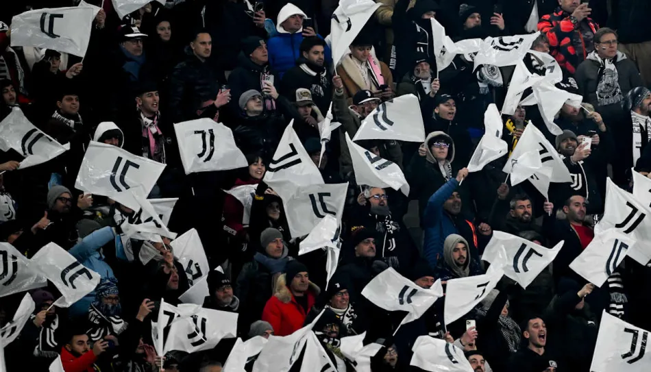 Tifosi della Juventus in uno stadio affollato sventolano bandiere bianche con il logo ufficiale del club, creando un'atmosfera carica di entusiasmo durante una partita. Supporter in abbigliamento invernale esultano e mostrano il loro sostegno alla squadra bianconera.