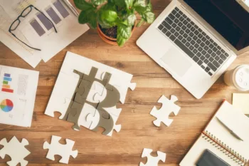 Su una scrivania in legno chiaro, sono poggiati un computer e dei documenti e un puzzle con vari pezzi composti e scomposti, a formare il logo del Bitcoin