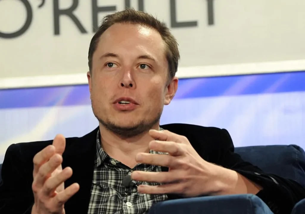 Elon Musk seduto durante una conferenza. Indossa una giacca blu scuro e camicia a scacchi