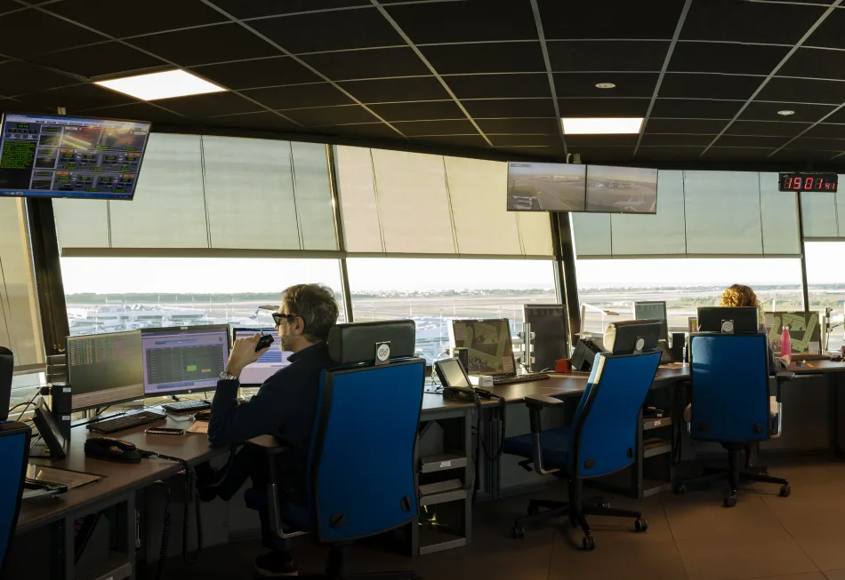 Torre di controllo dell'aeroporto con controllori di volo al lavoro su monitor e strumenti di navigazione aerea. Vista panoramica sulla pista di atterraggio e sui movimenti degli aerei.