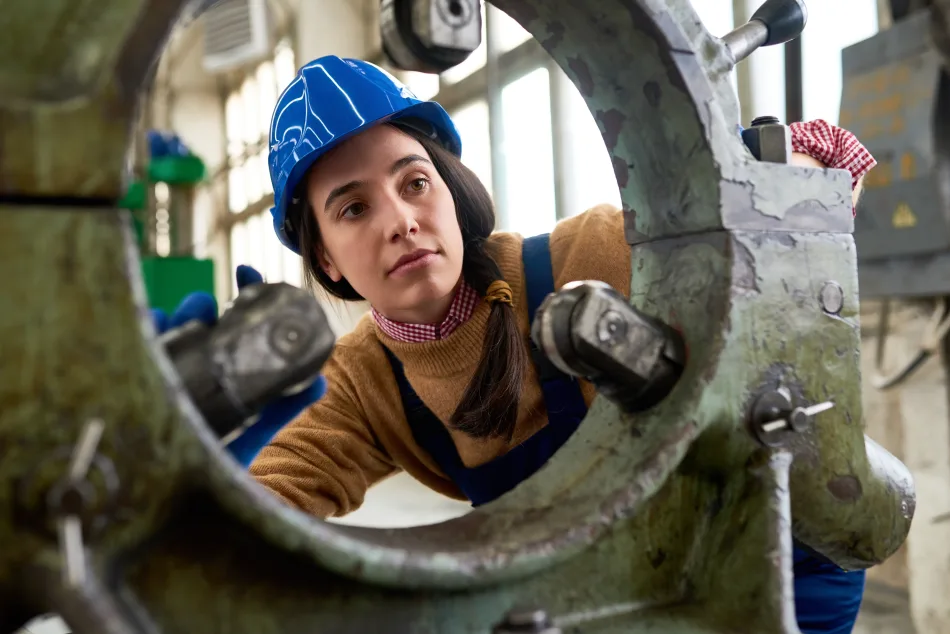 Donna meccanico al lavoro in fabbrica, indossando elmetto e tuta da lavoro, durante l'ispezione di una macchina.