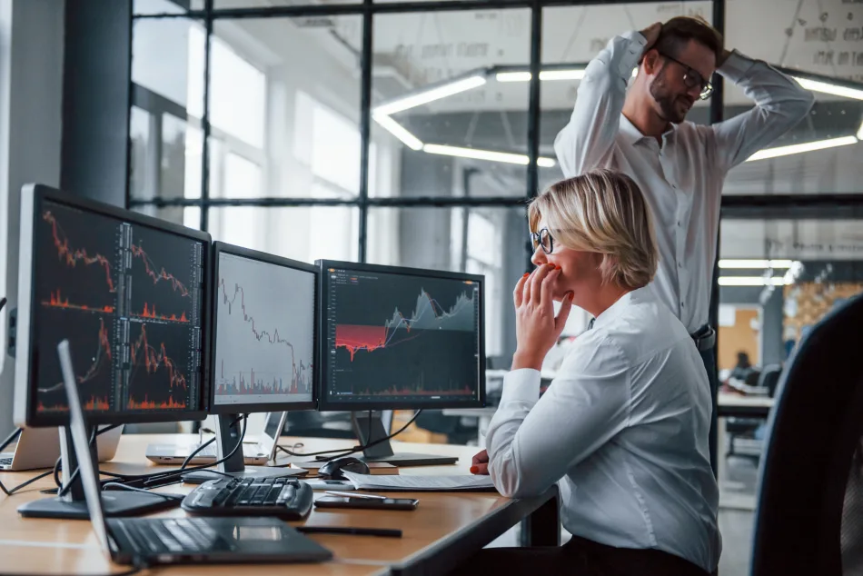 In un ufficio di una società di trading una donna osserva perplessa i grafici sui sei monitor che le stanno davanti mentre, alle sue spalle, un uomo in piedi, si mette la mani nei capelli