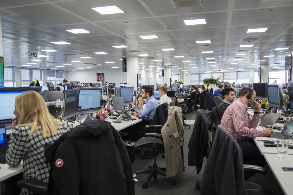 Sala trading con operatori finanziari al lavoro presso IG, tra computer, monitor e telefoni, in un ambiente moderno e tecnologico.
