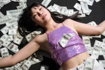 Donna asiatica distesa su un letto di banconote da cento dollari americani, in top glitterato viola.