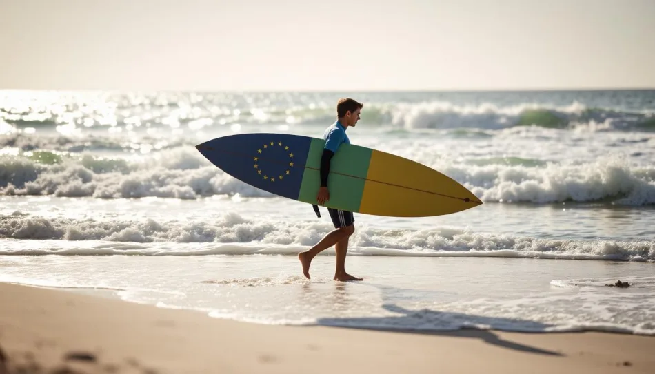 Giovane surfista con tavola da surf nei colori della bandiera dell’Unione Europea cammina sulla spiaggia verso il mare.