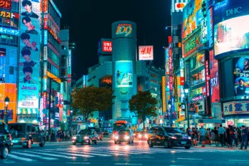 Strada notturna illuminata a Shibuya, Tokyo, con insegne al neon, traffico urbano e folla di pedoni.