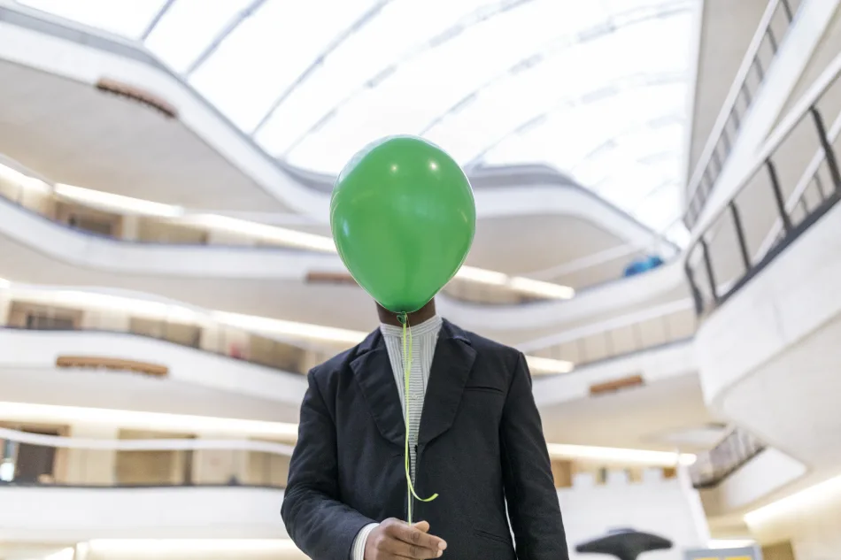 Uomo d’affari con volto nascosto da un palloncino verde in un edificio moderno, concetto di anonimato o incertezza finanziaria.