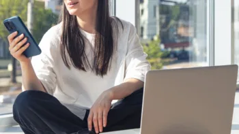 Una giovane donna allo smartphone davanti allo schermo del laptop