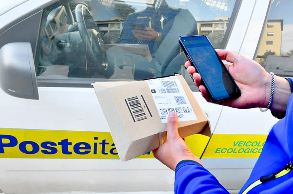 Foto ravvicinata che mostra un dipendente di Poste Italiane con in mano uno smartphone, con cui inquadra il codice a barre di un pacco prima di salire in automobile per una consegna.
