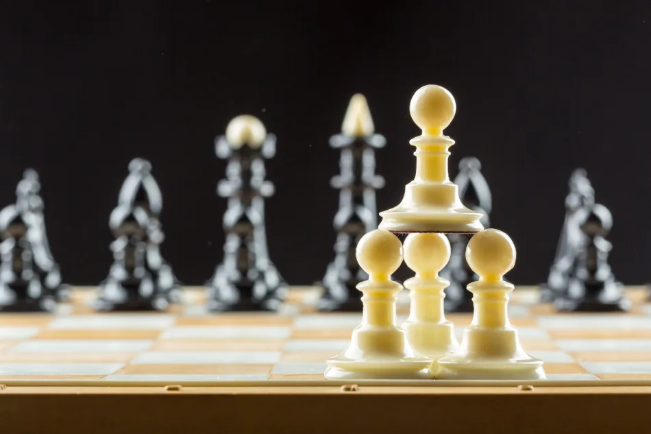 Gioco degli scacchi: tre pedine bianche sostengono un'altra pedina dello stesso colore, formando una torre altra più del re e della regina avversari, di colore nero, visibili sullo sfondo della scacchiera, leggermente sfuocati