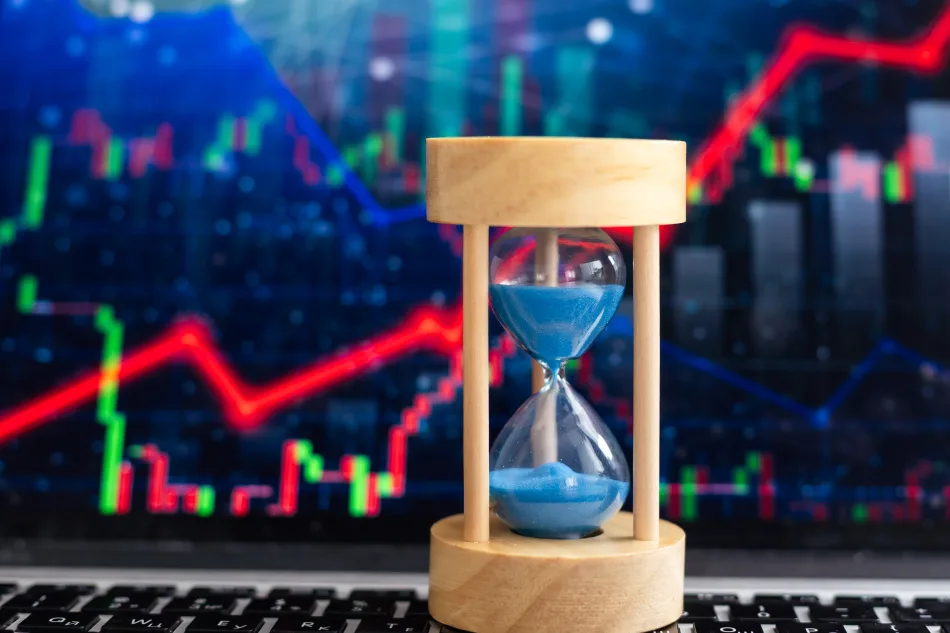 Una clessidra posizionata davanti a uno schermo che mostra dei grafici di attività finanziarie