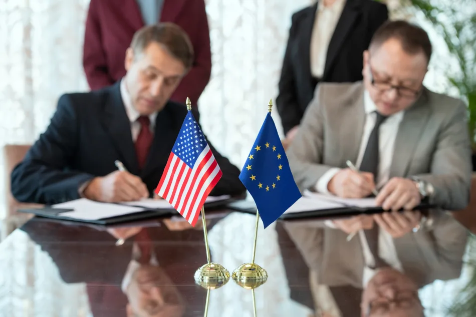 In primo piano su un tavolo le bandiere di Stati Uniti e Unione europea mentre, sullo sfondo, due persone firmano dei documenti
