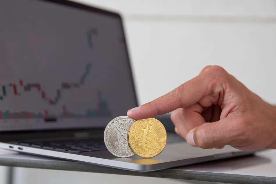 Ether e Bitcoim davanti a un laptop che mostra un grafico a candele giapponesi