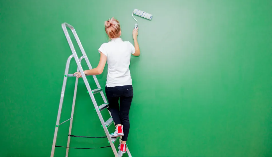 Una donna, su una scala, ha in mano un roller con cui sta dipingendo di verde un muro