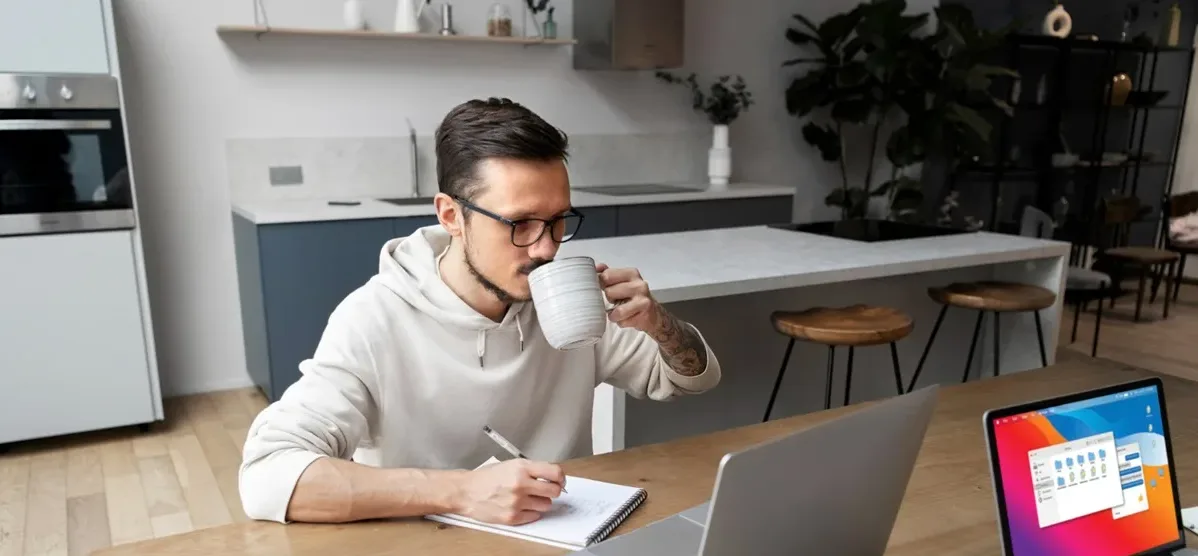 Un giovane lavoratore in smart working. Seduto al tavolo della cucina con il pc, il tablet e un blocchetto appunti, segue una riunione mentre beve una tazza di te